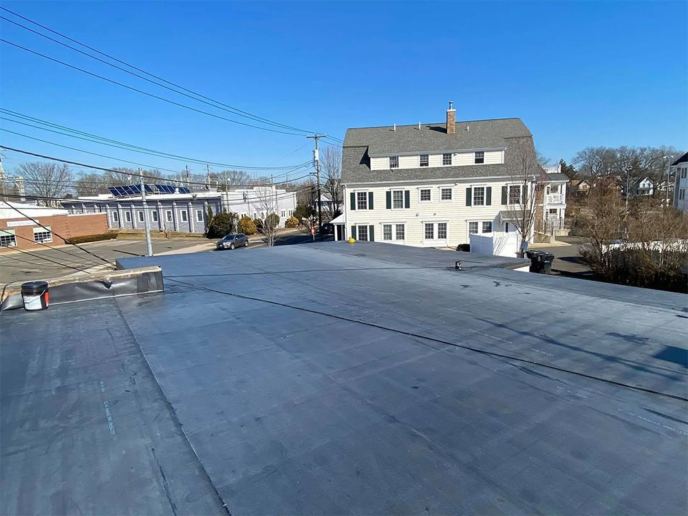A wide shot of a flat, gray roof. A white building with a gray roof stands in the background, under a blue sky.