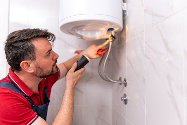Plumber inspecting a water heater, using a flashlight in a tiled bathroom.
