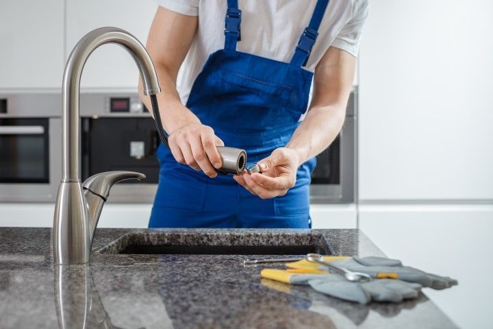 Plumber in blue overalls working on faucet in kitchen.