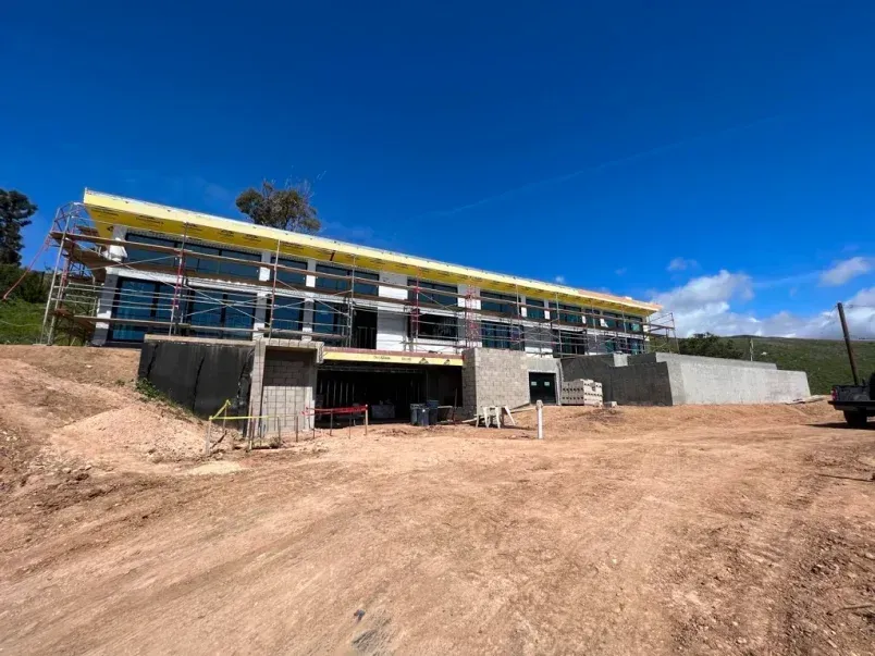 Two-story building under construction on a hillside, blue sky, scaffolding, dirt road in front.
