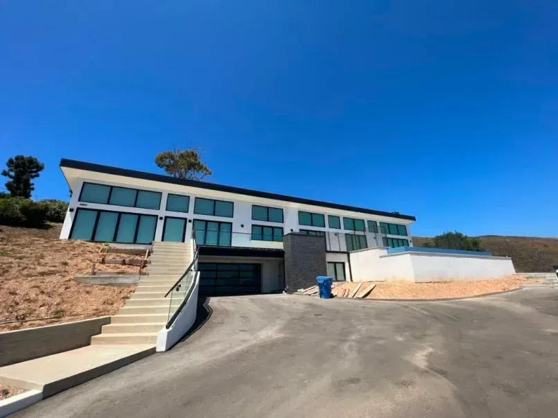 Modern white house with black trim and large windows, on a hill with a blue sky.