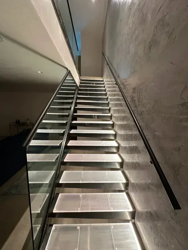 Modern staircase with illuminated steps, glass railing on one side, and dark handrail on the other.