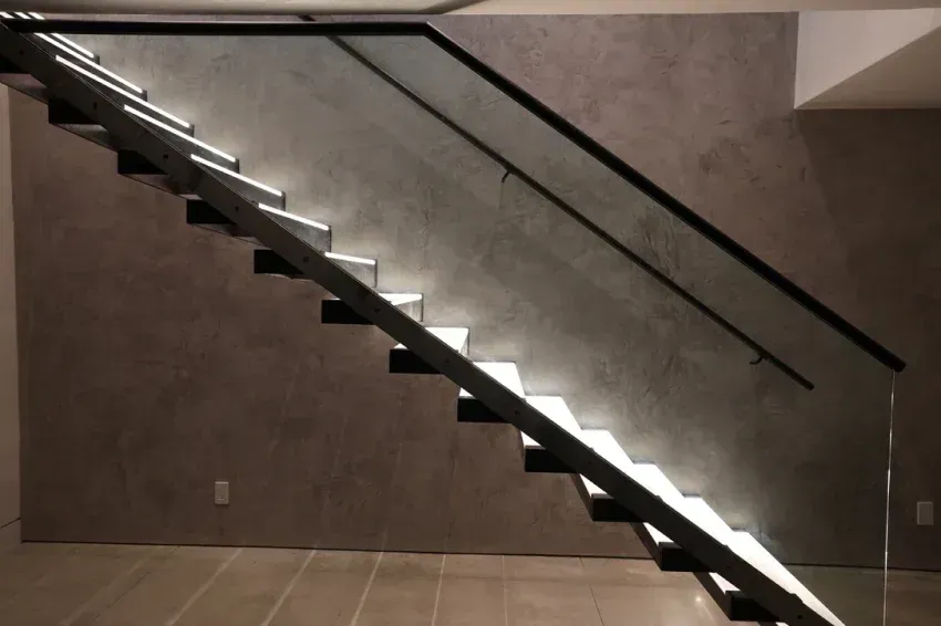 Modern staircase with lit steps, glass railing, and textured gray wall.