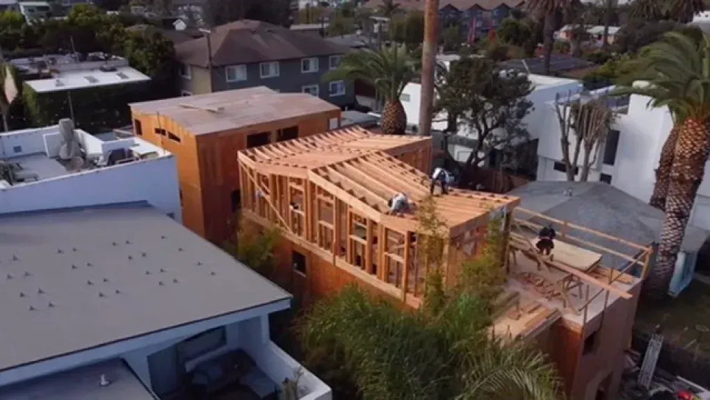 Construction site: wooden building frame with workers on the roof, surrounded by houses and palm trees.