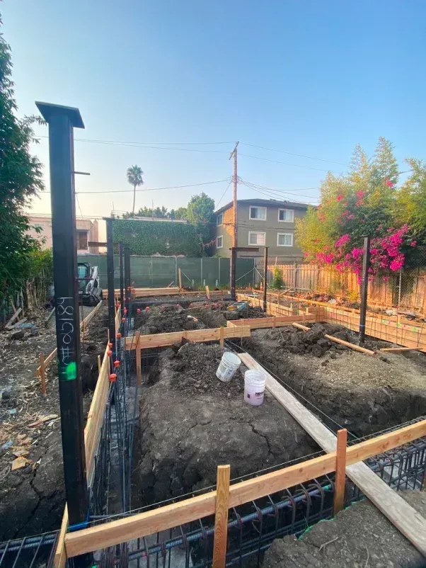 Construction site with wooden forms, rebar, dirt, and metal support beams. Building exterior under construction.