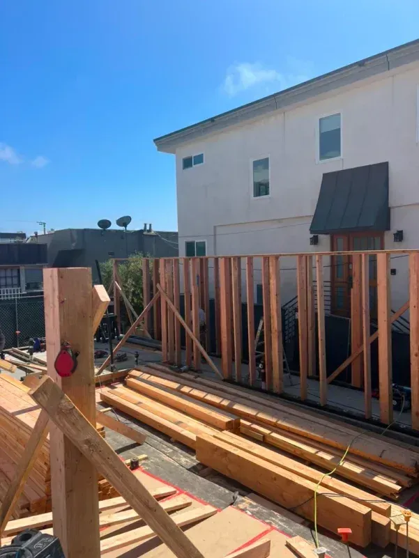Construction site with wooden framework, building materials, and a two-story building against a blue sky.