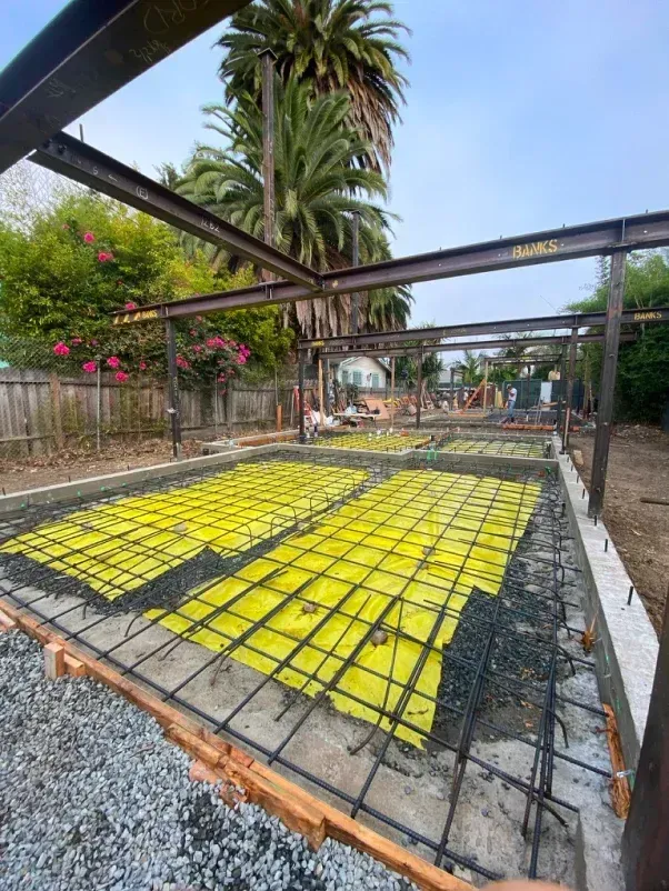 Construction site with steel frame, rebar, gravel, and yellow sheeting, outdoors.