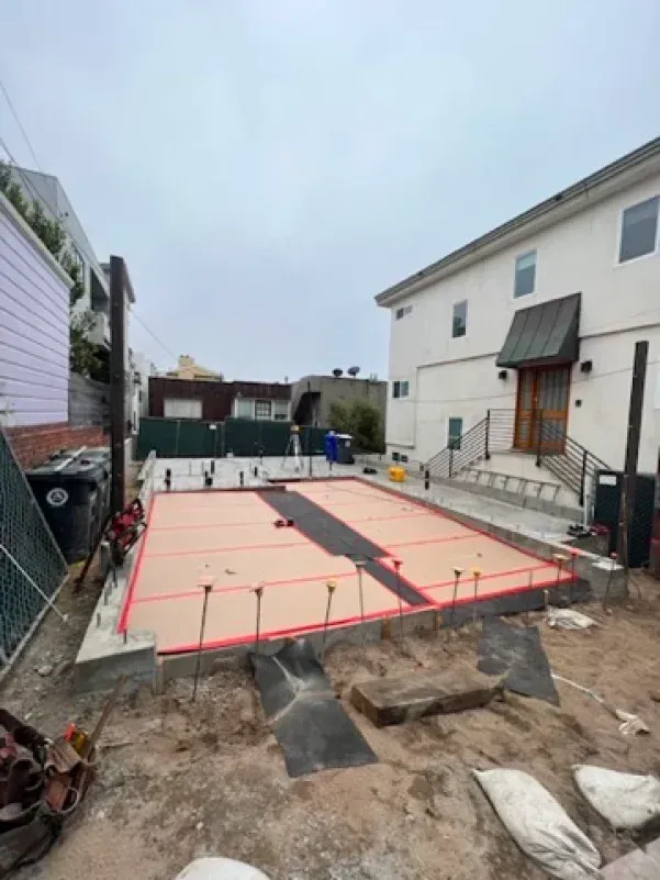 Construction site: a concrete slab framed with orange barriers. Buildings and sky visible in the background.
