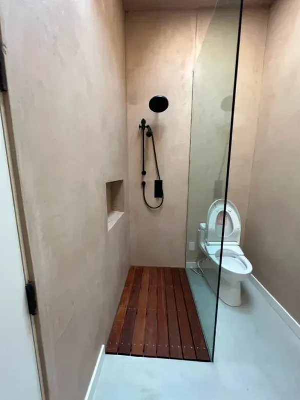 Small, beige-walled bathroom with glass shower, toilet, and wooden shower floor. Black shower fixtures.