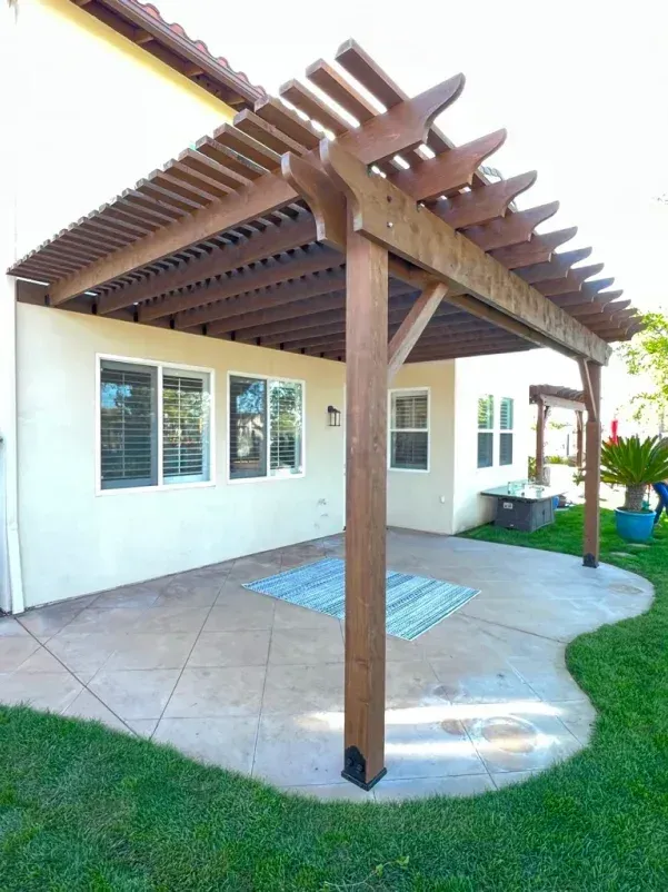 Wooden pergola covering a patio with a rug. The patio is beside a beige building with windows. Green grass surrounds it.