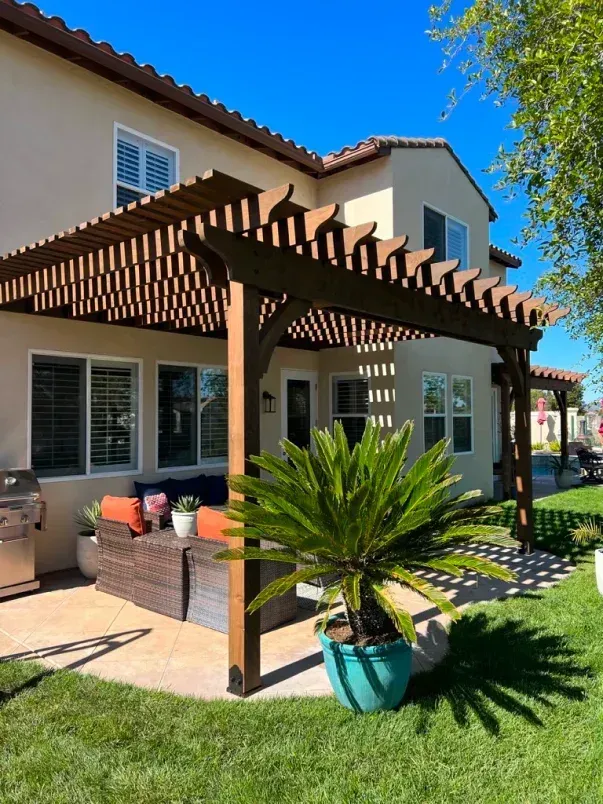 Pergola shades patio with seating in front of two-story house. Palm tree in turquoise pot sits on patio.