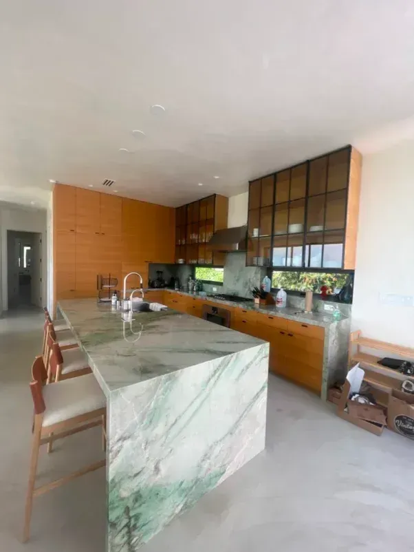 Modern kitchen with green marble island, light wood cabinets, glass-front uppers, and bar stools.