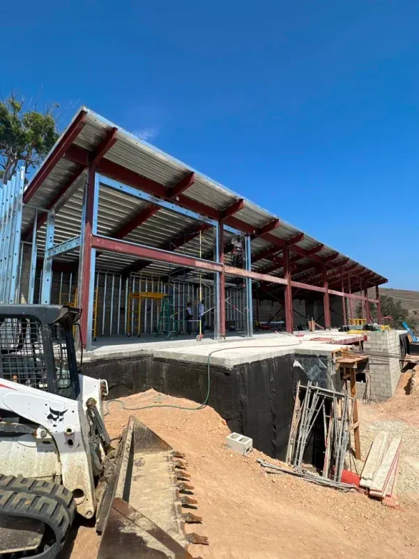 Construction site with steel frame building and a Bobcat in front, blue sky.