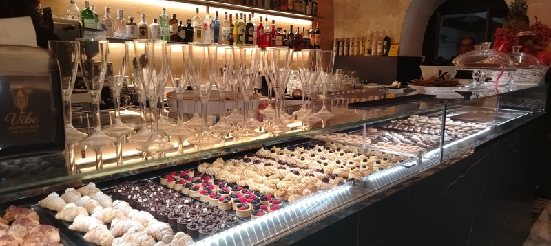 Una vetrina piena di pasticcini e crostatine di vario genere in un caffè scarsamente illuminato, con un bancone retroilluminato e dei bicchieri sopra.