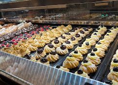 File di pasticcini ripieni di crema con ciliegie e frutti di bosco freschi, disposti su vassoi all'interno di una vetrina da pasticceria.