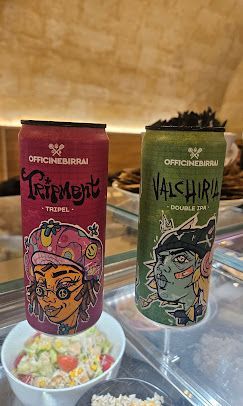 Due lattine di birra Officina della Birra, una rossa Tripment e una verde Valchiria, appoggiate su una superficie di vetro sopra del cibo.