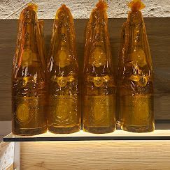 Quattro bottiglie di champagne avvolte in una lamina dorata traslucida, allineate su uno scaffale illuminato.