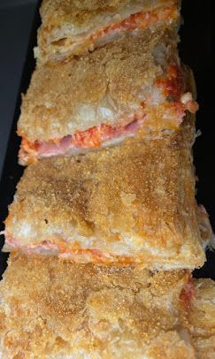 Una pila verticale di snack fritti, impanati e dorati, ripieni di formaggio fuso e prosciutto, adagiata su una superficie scura.