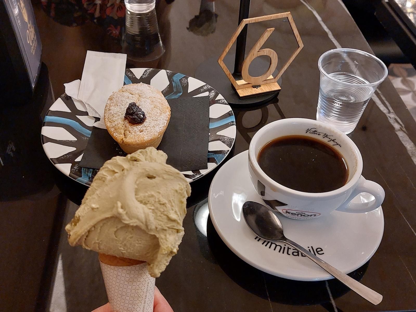Una mano che regge un cono gelato al pistacchio accanto a una piccola crostata e una tazza di caffè su un tavolo con il numero sei.