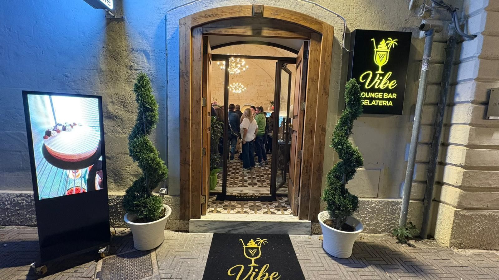 L'ingresso di Vibe, un lounge bar con un portale ad arco in legno, piante ornamentali a spirale, tappeti e insegne con il logo del locale.