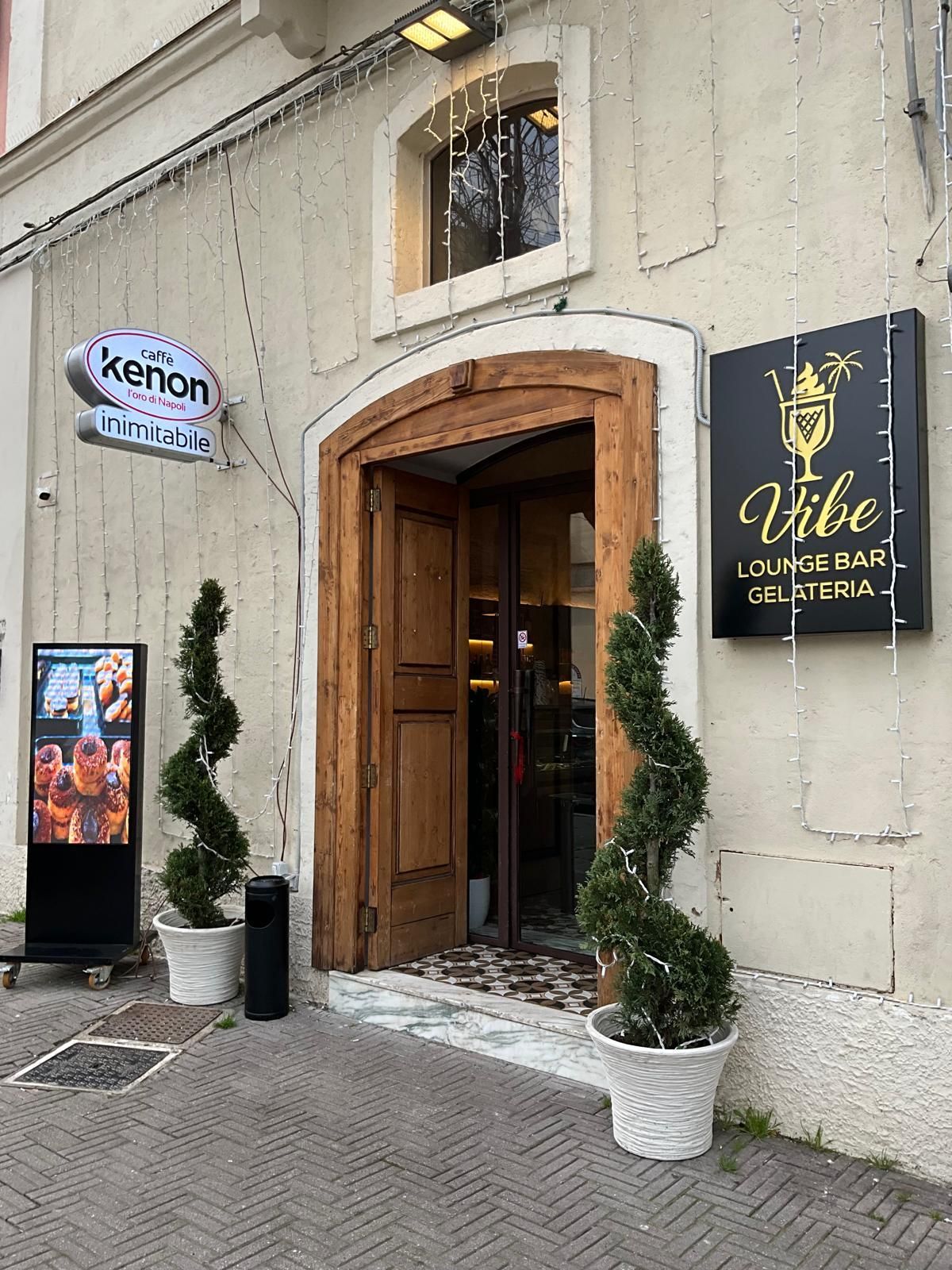Vista esterna dell'ingresso del lounge bar e gelateria Vibe, con due piante in vaso a spirale che fiancheggiano una porta di legno.