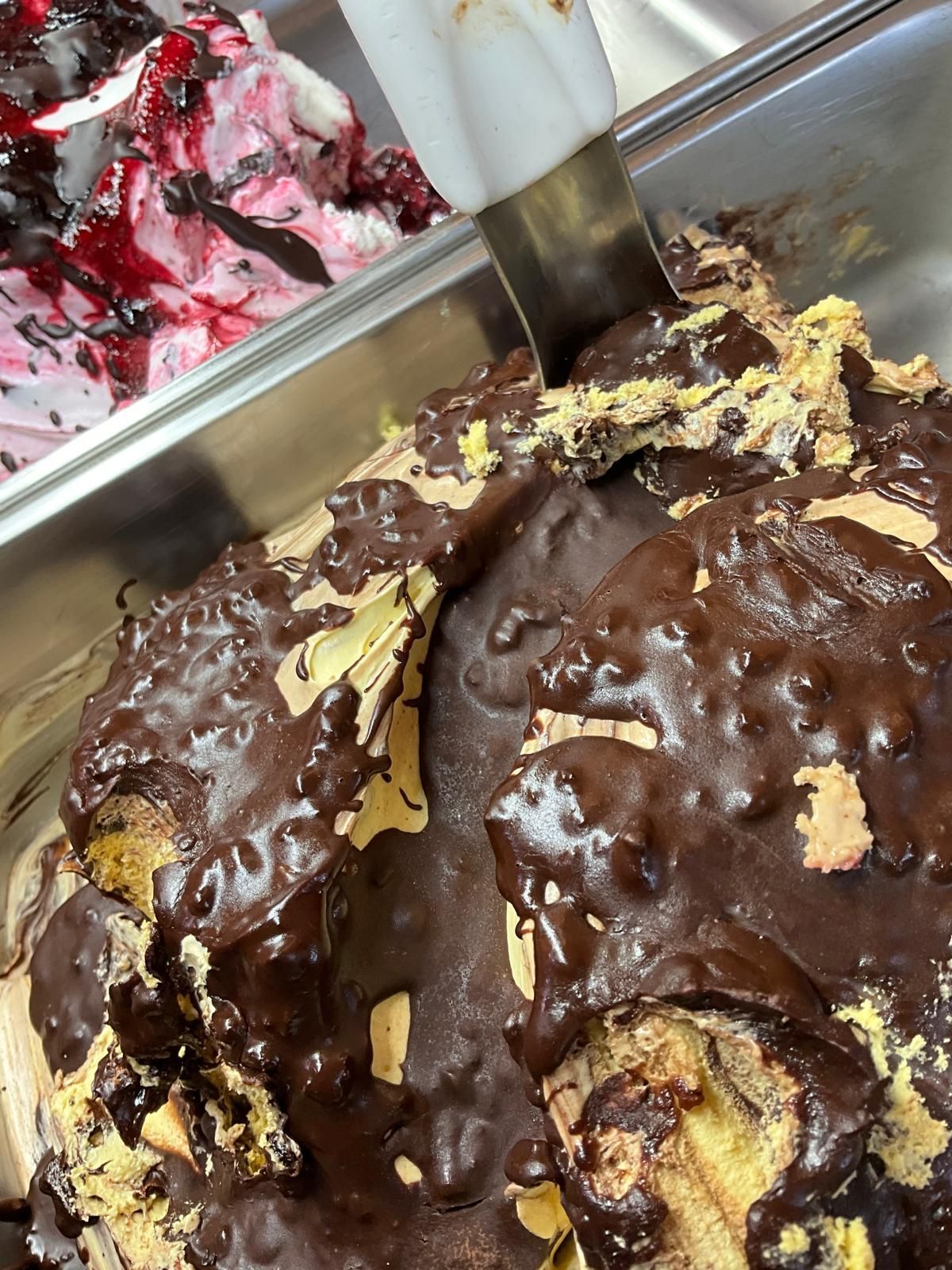 Un contenitore di metallo con palline di gelato giallo ricoperte da una spessa e lucida spirale di cioccolato, accanto a gelato ai frutti di bosco.