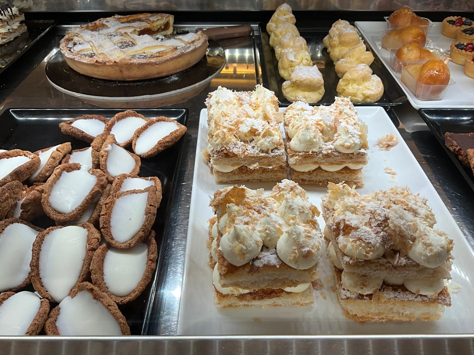 Una vetrina che espone vari tipi di pasticcini, tra cui crostatine alla crema, profiteroles, torte a strati con crema e dolcetti rotondi.
