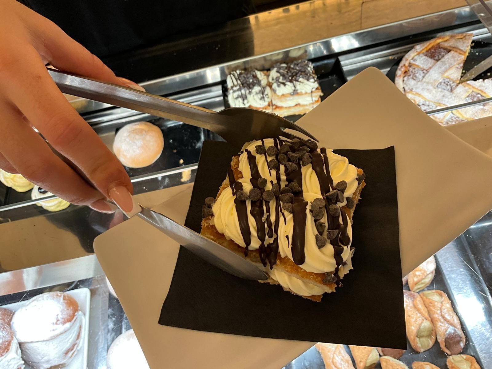 Una mano, usando delle pinze di metallo, solleva un pasticcino ripieno di crema, guarnito con una colata di cioccolato e scaglie di cioccolato, e lo adagia su un tovagliolo nero.