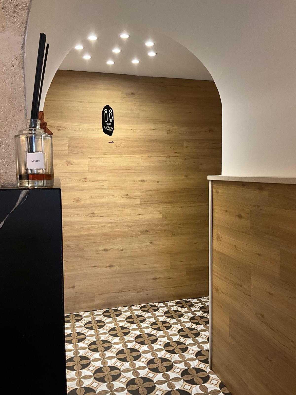 Un corridoio con una parete di fondo rivestita in legno, pavimento piastrellato con motivi decorativi e un cartello del bagno sotto un soffitto a volta.
