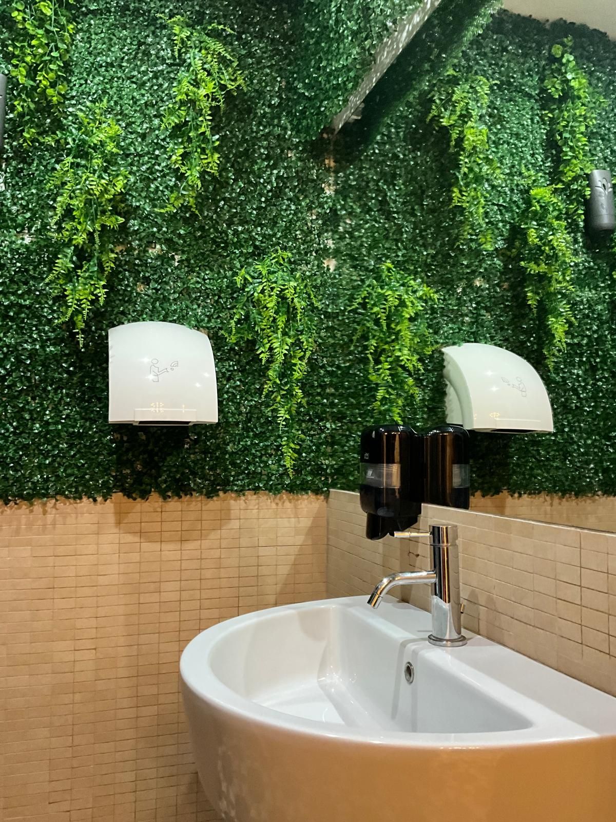 Un lavabo da bagno con dispenser di sapone e asciugamani elettrico, addossato a una parete piastrellata ricoperta di piante verdi artificiali sospese.