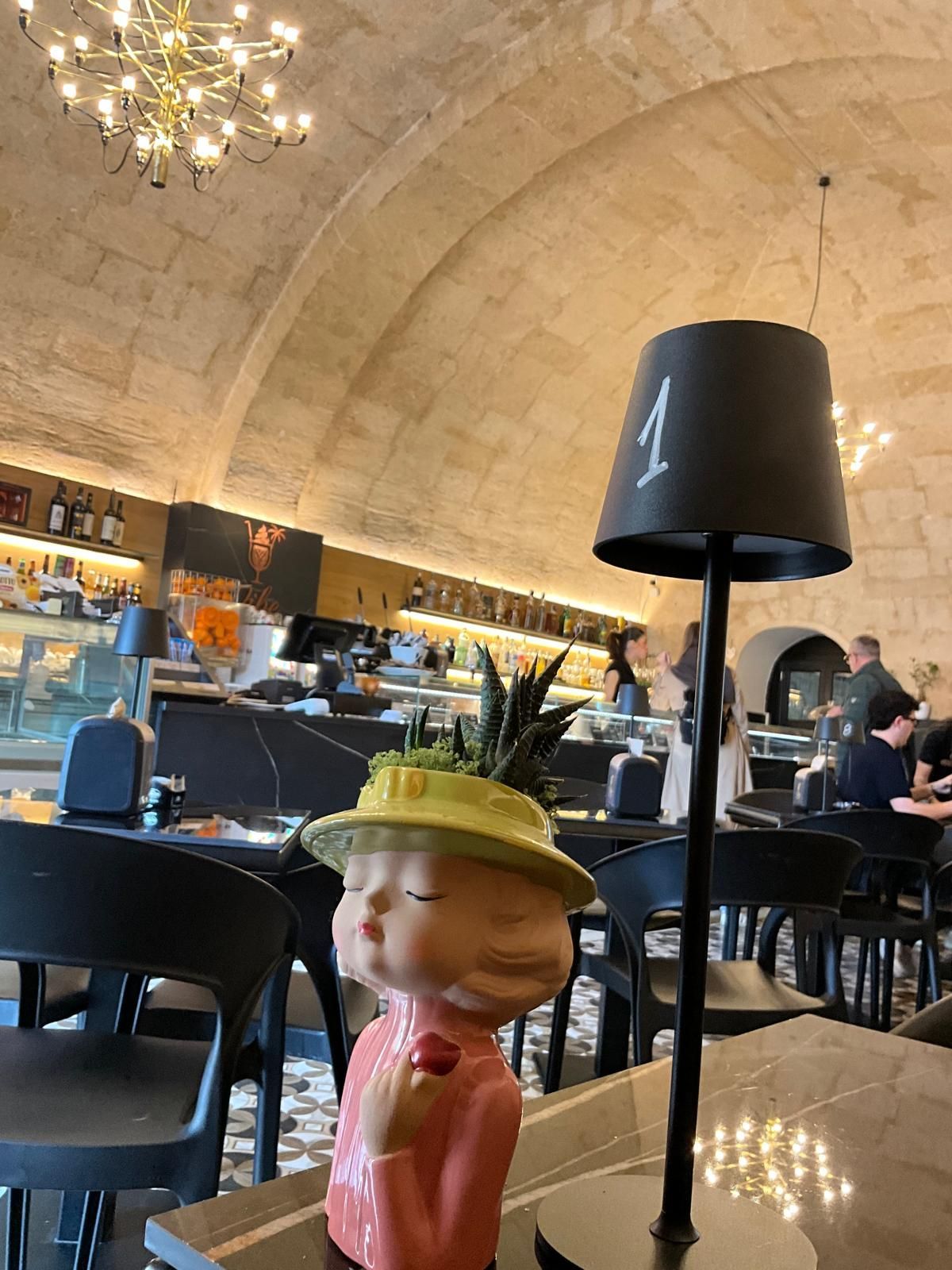 Una lampada da tavolo numero 1 si trova accanto a una fioriera decorativa a forma di persona con un cappello giallo in un caffè con archi in pietra.