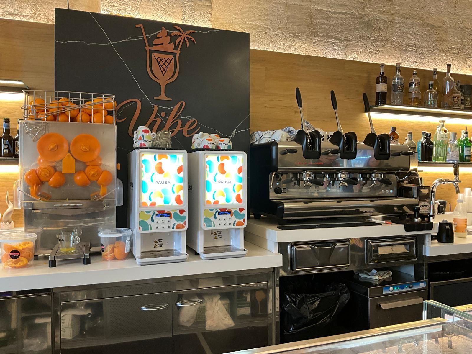 Un bancone bar con spremiagrumi, due distributori di bevande e una macchina per caffè espresso professionale, addossato a una parete in legno.
