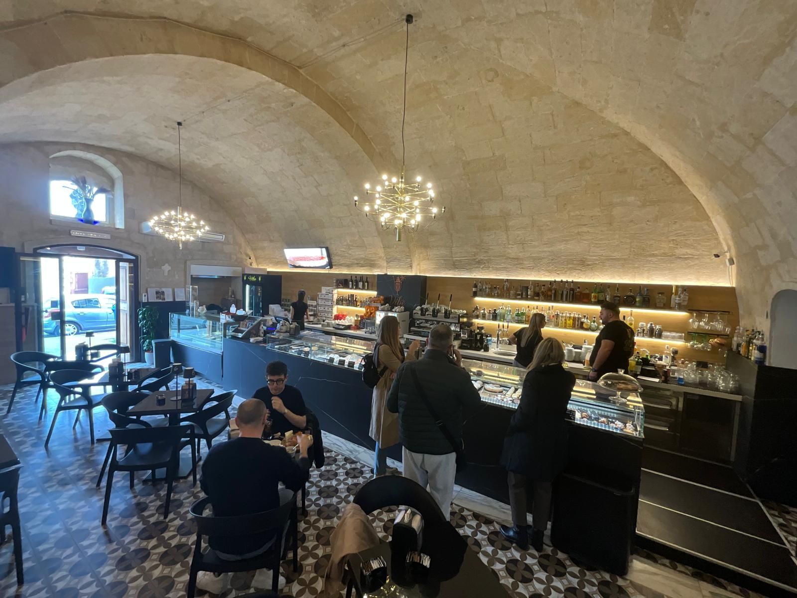 Un caffè con soffitto a volta, dove i clienti siedono ai tavoli e il bancone espone i prodotti alimentari sotto lampade a sospensione.