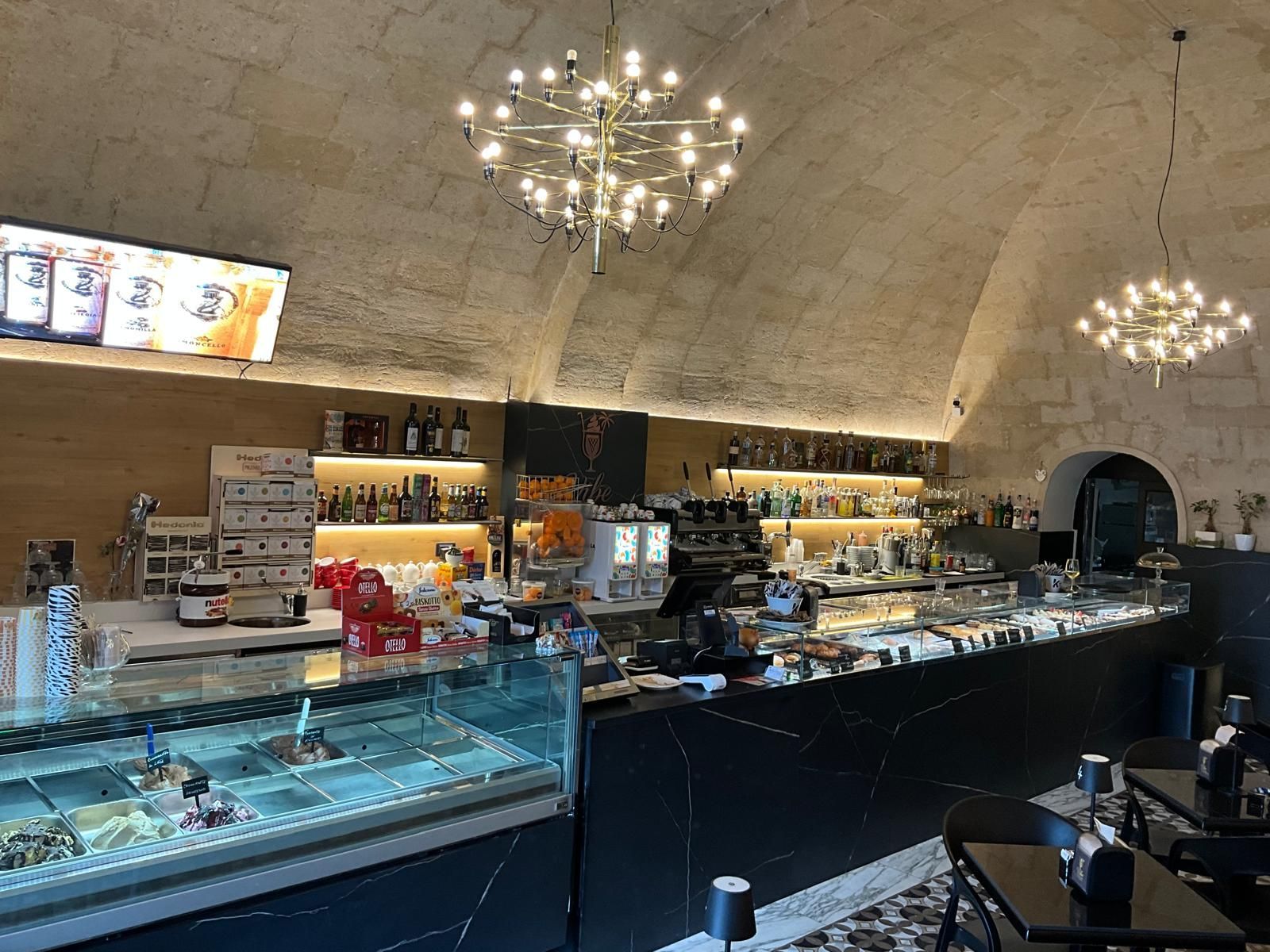 Vista interna di una caffetteria con soffitti a volta in pietra, due lampadari moderni, una vetrina per il gelato e un bancone scuro.