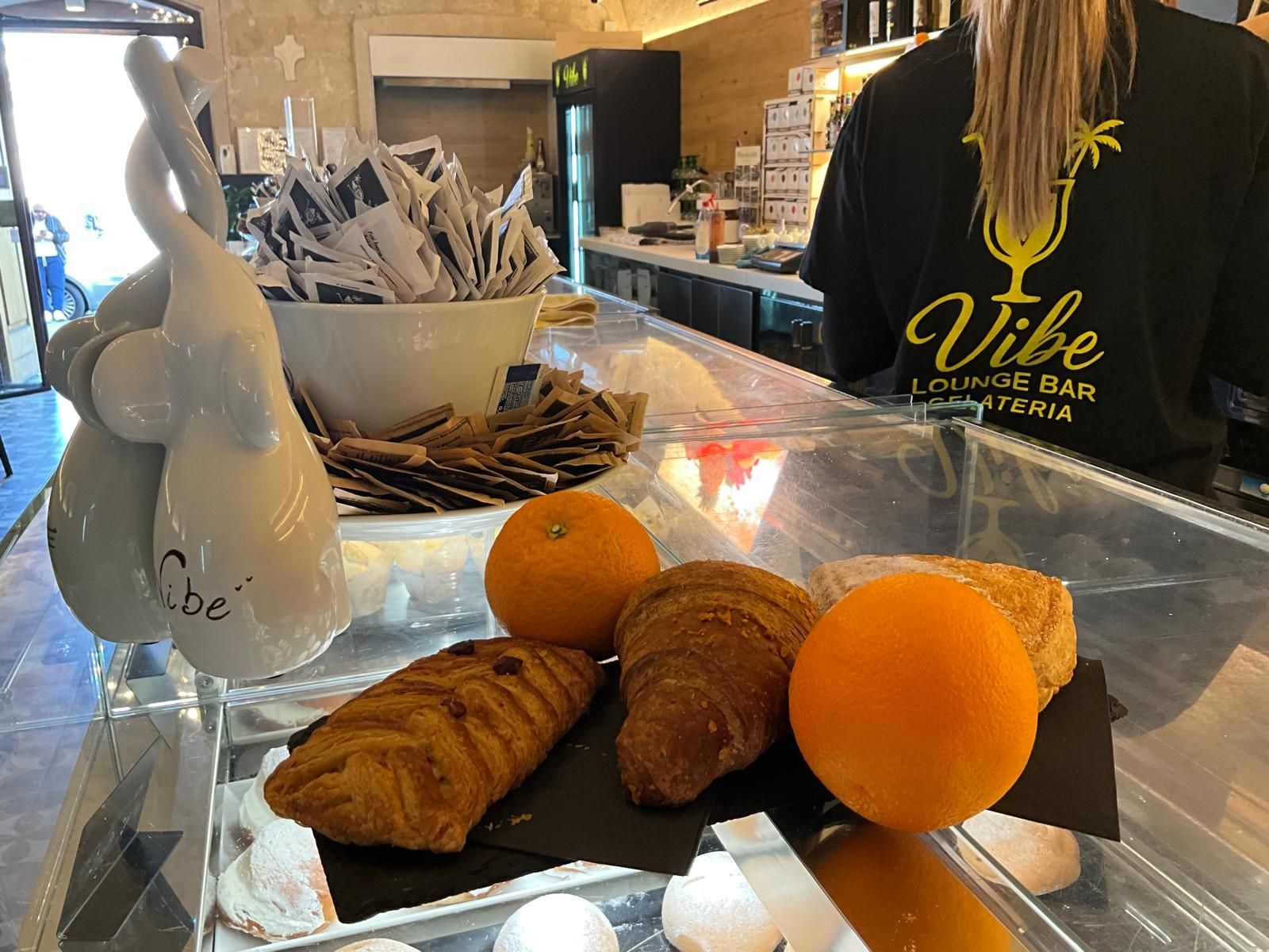 Una vetrina del Vibe Lounge Bar di Catania con croissant, pasticcini e arance intere su un bancone di vetro.