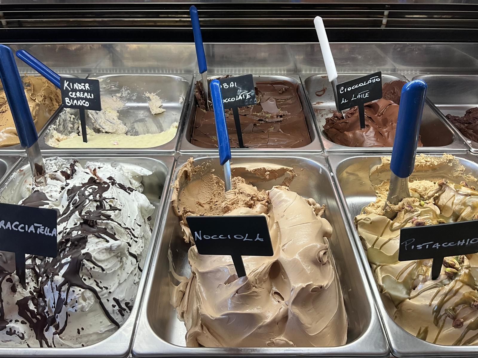 Una vetrina piena di gelati di vari gusti in contenitori di metallo, ognuno con un'etichetta nera e una pallina da gelato.