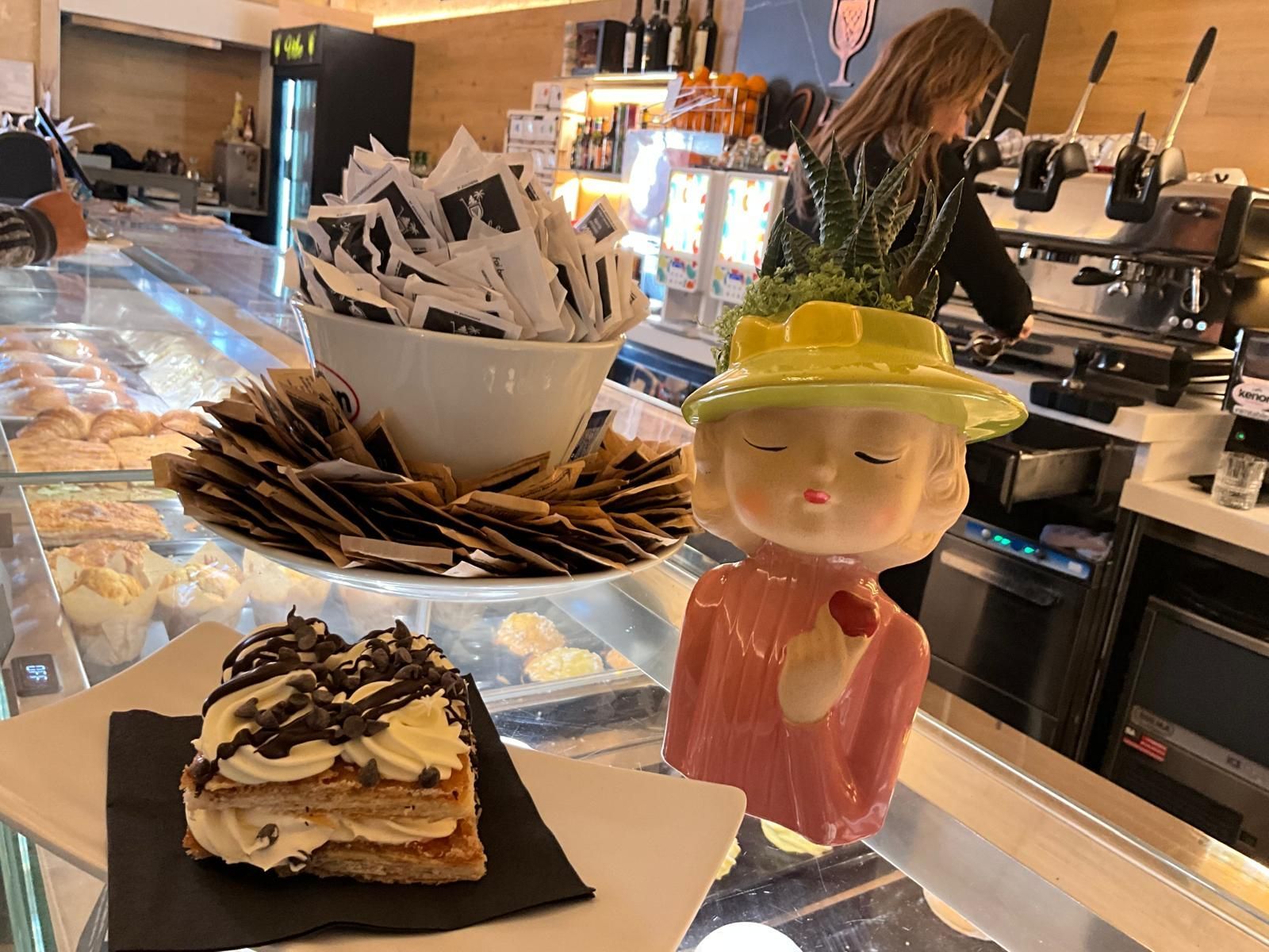 Un pasticcino con crema e cioccolato è appoggiato su un bancone accanto a un vaso decorativo a forma di busto e a una ciotola di bustine di zucchero.