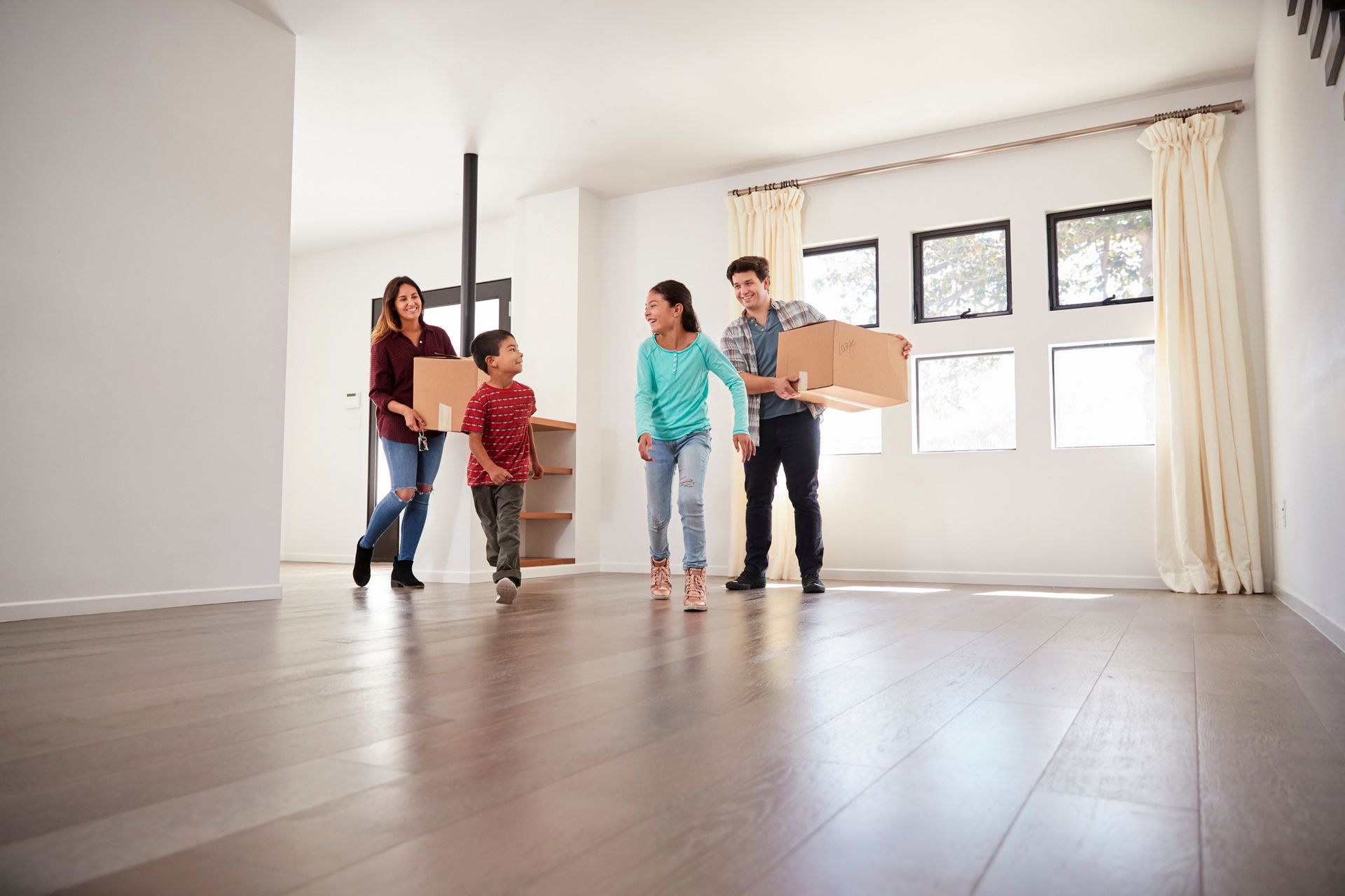 Family entering a brightly lit home, dog in front. Wooden floors, staircase, and side table with decor.