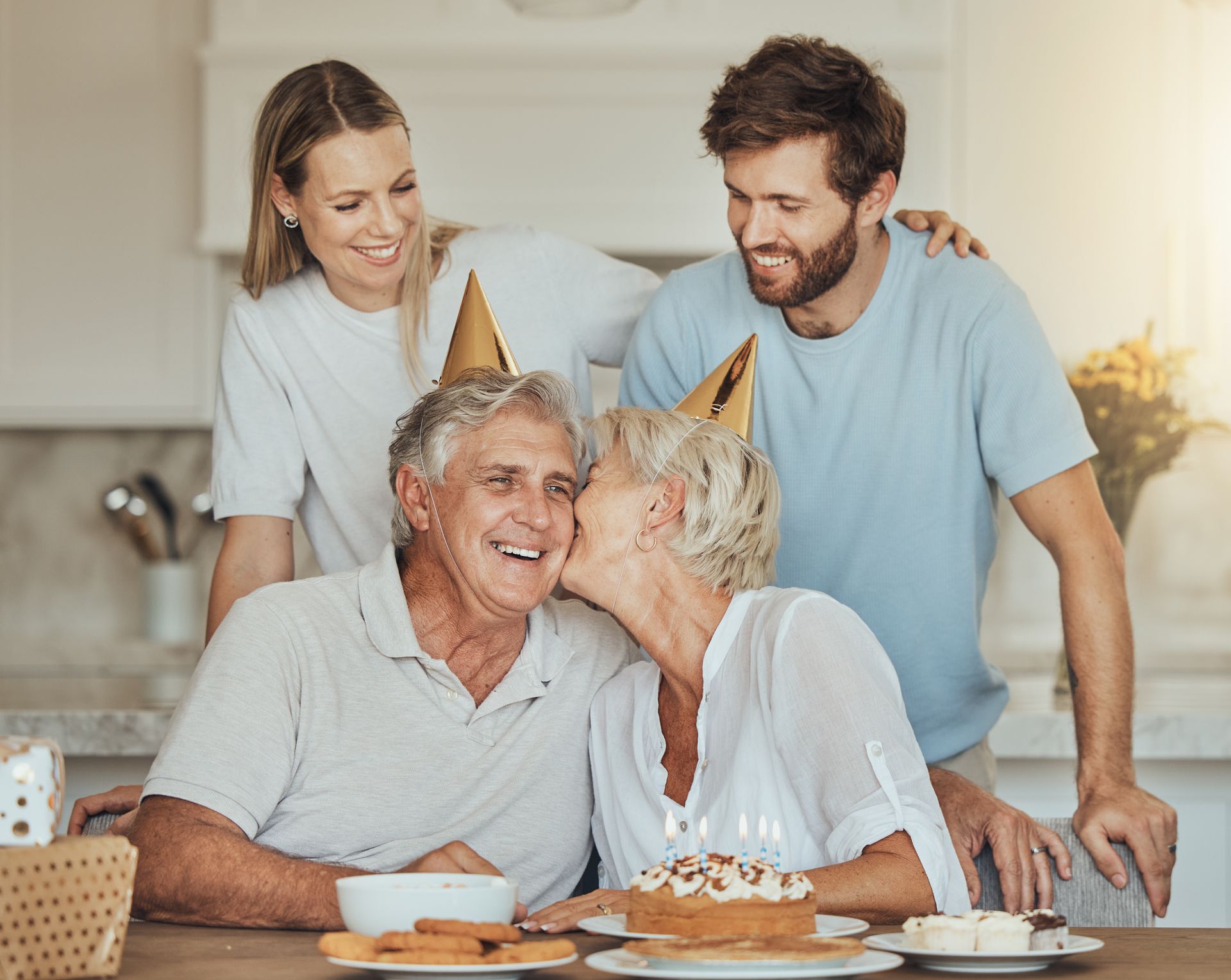 Family celebrating a birthday: grandparents with party hats, cake, being kissed, two adult children watching.