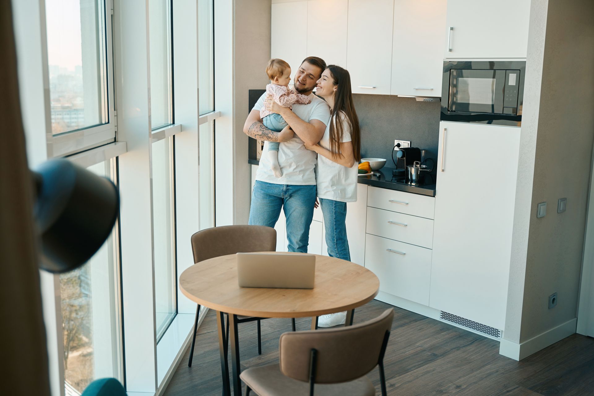 Family in a modern kitchen. Man holding a baby, woman smiling. Round table with laptop, large window.