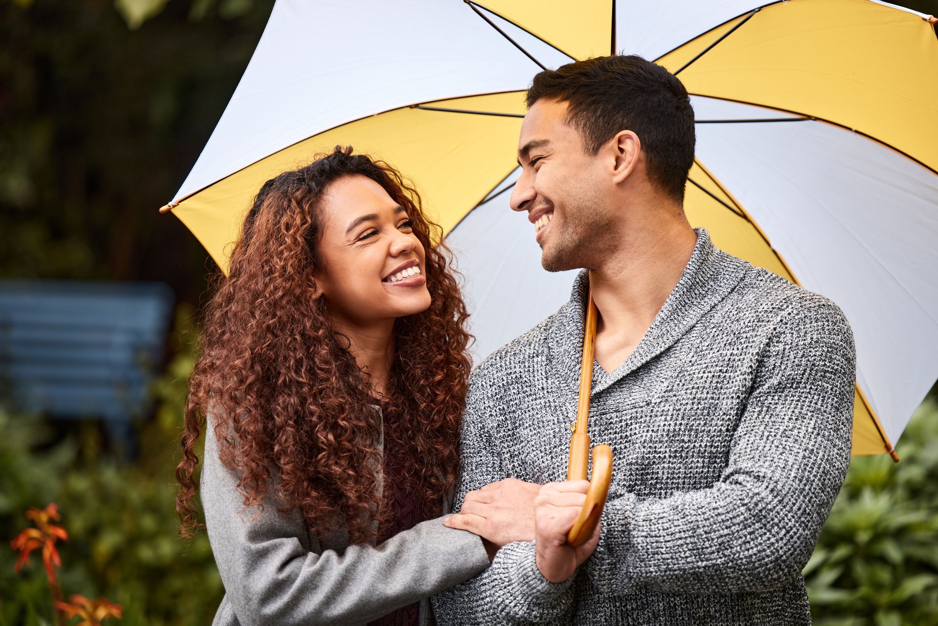 Smiling couple under a yellow and white umbrella, outdoors in a garden setting.