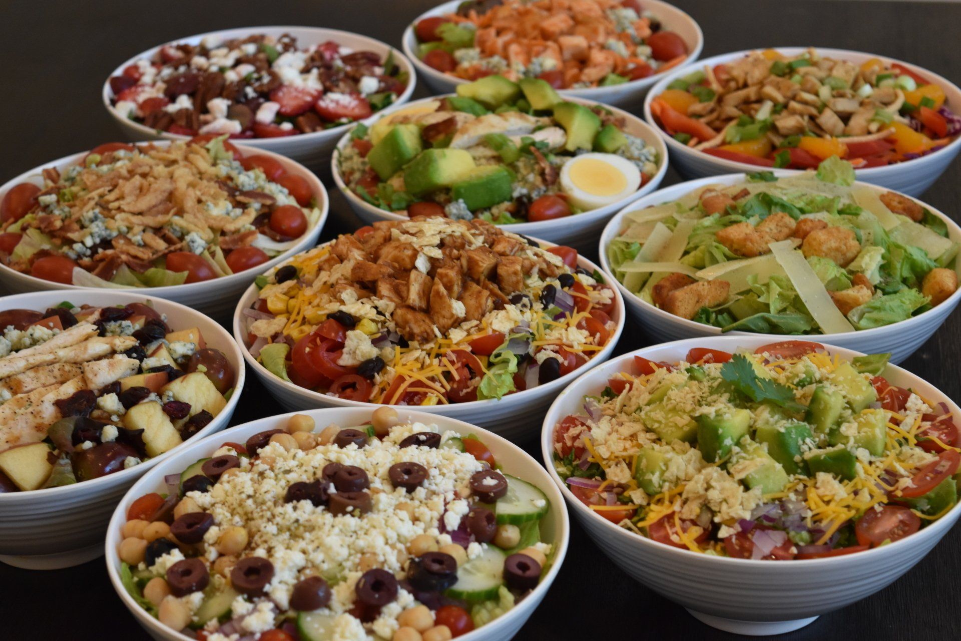 Assorted salads in white bowls, featuring various toppings like avocado, cheese, and nuts.