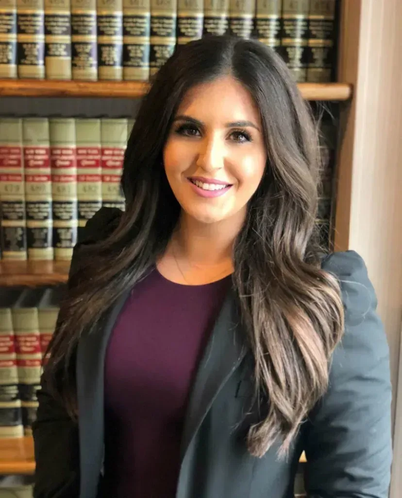Woman with long dark hair, smiling, wearing a blazer and purple top in front of bookshelves.