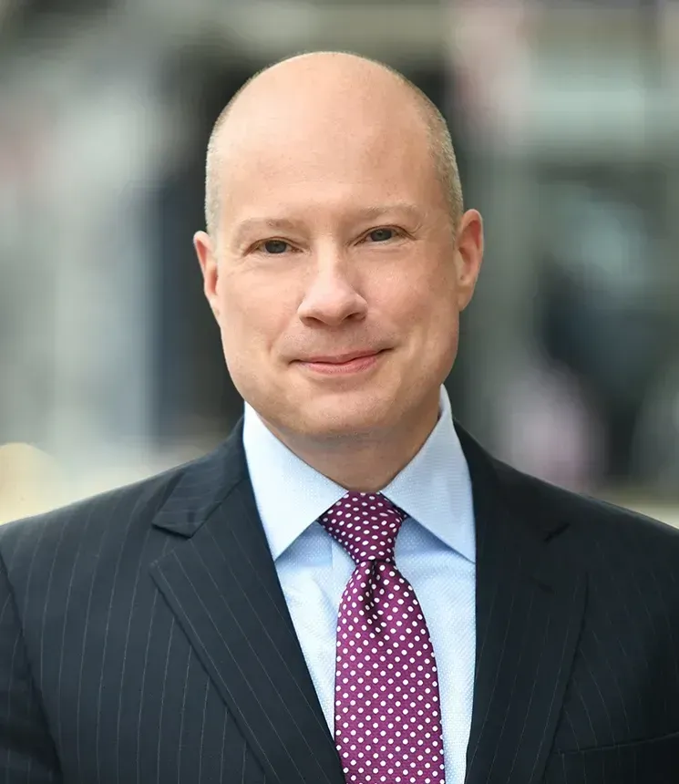 Bald man in suit and purple tie, smiling, blurred building background.