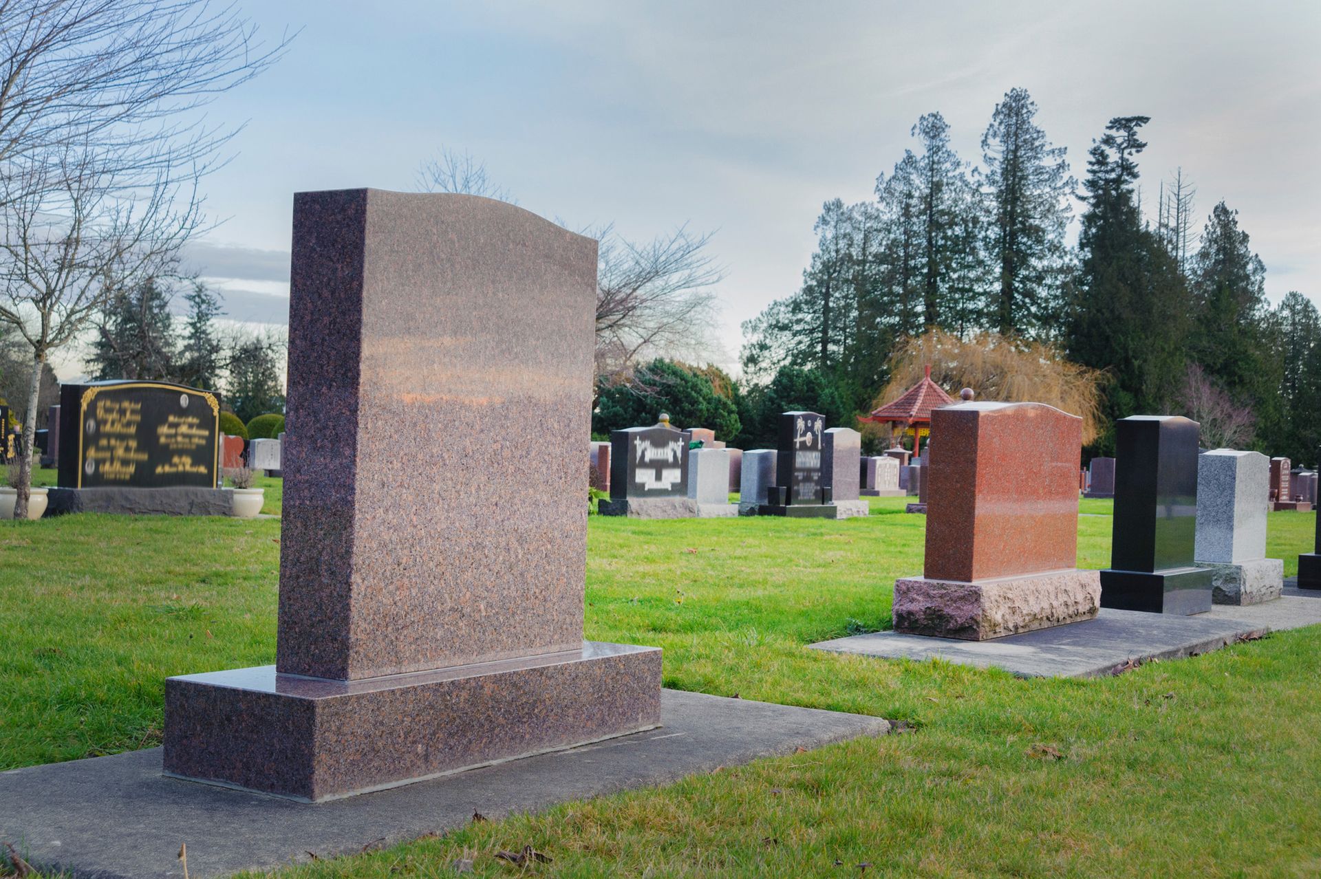 Gravestone in a cemetery, with green grass and trees under a cloudy sky.