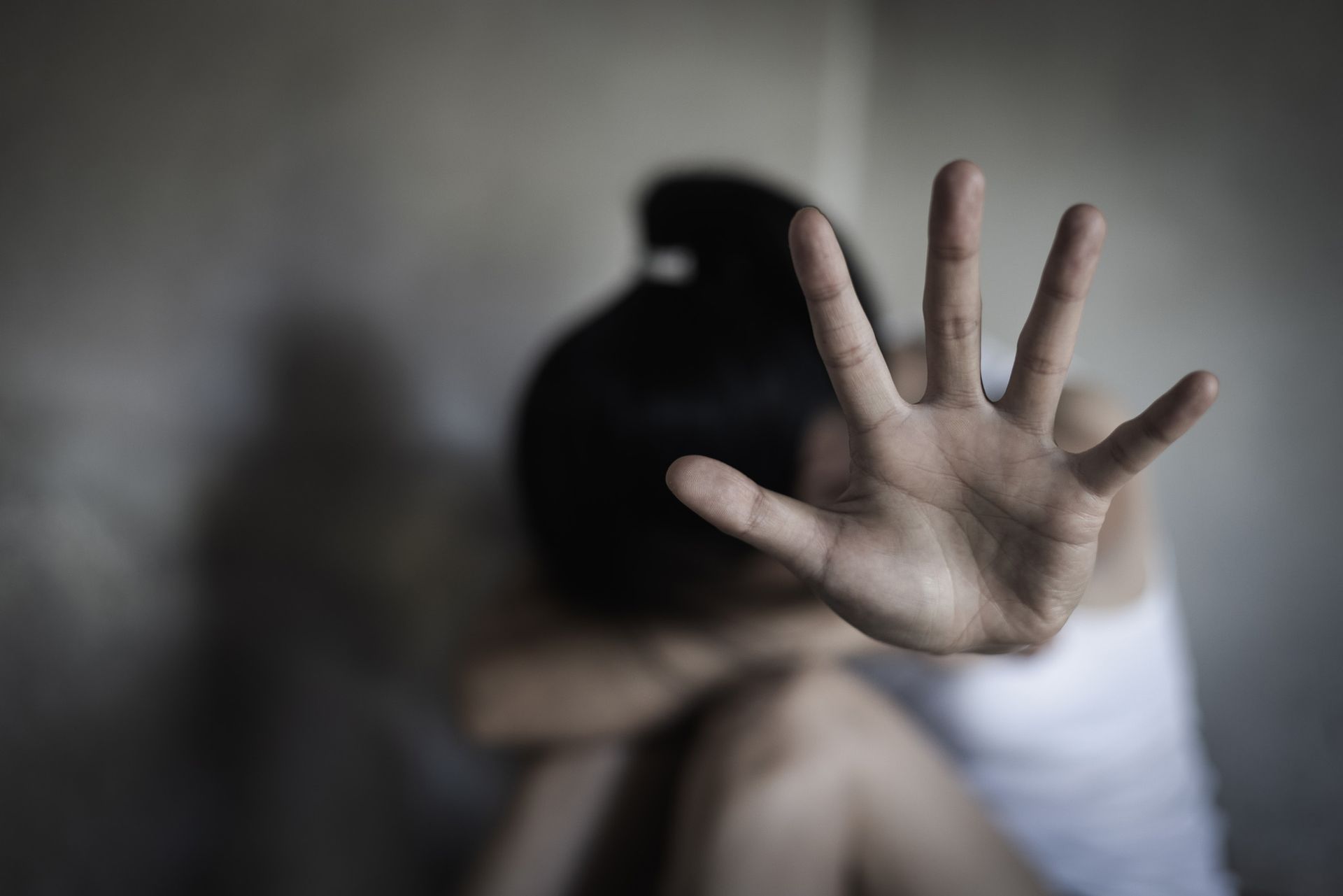 Woman raising hand to stop, hiding her face, in a dimly lit room, appears to be in distress.
