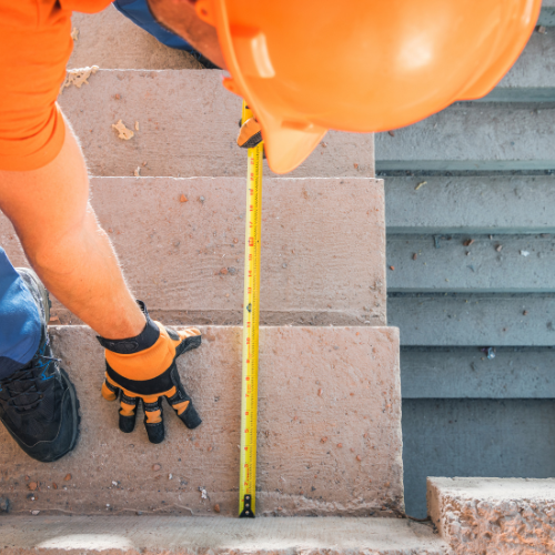Person in orange hard hat and gloves measures a step with a yellow tape measure.