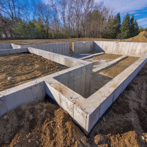 Concrete foundation of a building under construction, on a dirt lot, with trees in the background.