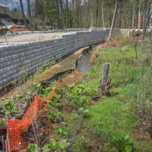 Stone retaining wall bordering a stream, with orange safety fencing and vegetation in the foreground.