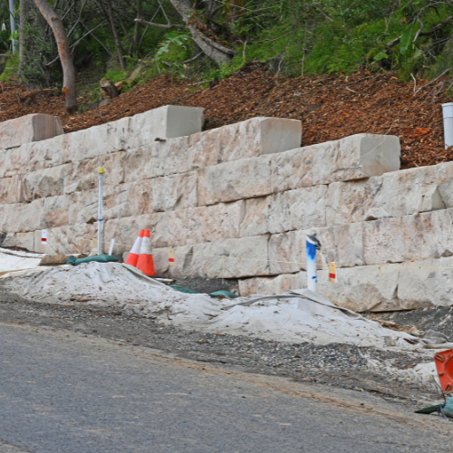 Stone retaining wall on a hillside, with construction materials and orange cones, next to a road.
